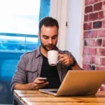 young-man-sitting-in-front-of-his-laptop-with-a-cup-of-coffee-in-hand-while-texting-photo