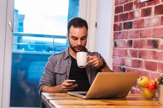 young-man-sitting-in-front-of-his-laptop-with-a-cup-of-coffee-in-hand-while-texting-photo